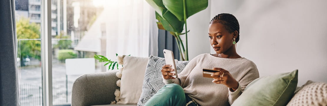 Woman sits on a couch with her credit card and her cell phone.